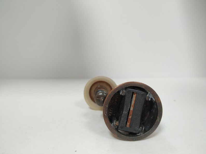 May include: Close-up of a vintage pencil sharpener with a bronze-colored metal base and a clear, cylindrical plastic top. The sharpener has a blade visible inside and is set against a white background.