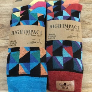 Könnte beinhalten: Zwei Paar bunte Socken mit geometrischen Mustern. Die Socken zeigen eine Mischung aus blauen, orangefarbenen, lila und schwarzen Dreiecken. Jedes Paar ist mit "HIGH IMPACT cotton rich" und Größenangaben versehen. Auf einer Holzoberfläche.