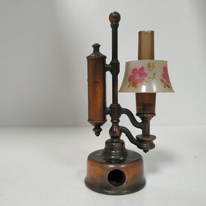 May include: Antique bronze-colored pencil sharpener shaped like a lamp. It features a cylindrical body, a floral-patterned lampshade, and a pencil entry hole. The lamp-shaped sharpener is a unique desk accessory.