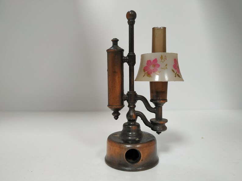 May include: Antique bronze-colored pencil sharpener shaped like a lamp. It features a cylindrical body, a floral-patterned lampshade, and a pencil entry hole. The lamp-shaped sharpener is a unique desk accessory.