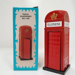 May include: Red telephone box shaped pencil sharpener with a gold crown on top. The sharpener has the word "Telephone" written on the front. The box is blue with a white border and says "Pencil Sharpener No. 170A".