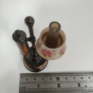 May include: Miniature bronze-colored desk lamp with a floral patterned lampshade. The lamp has a small base and a slender stem. The lampshade is decorated with pink flowers. A ruler is placed next to the lamp.