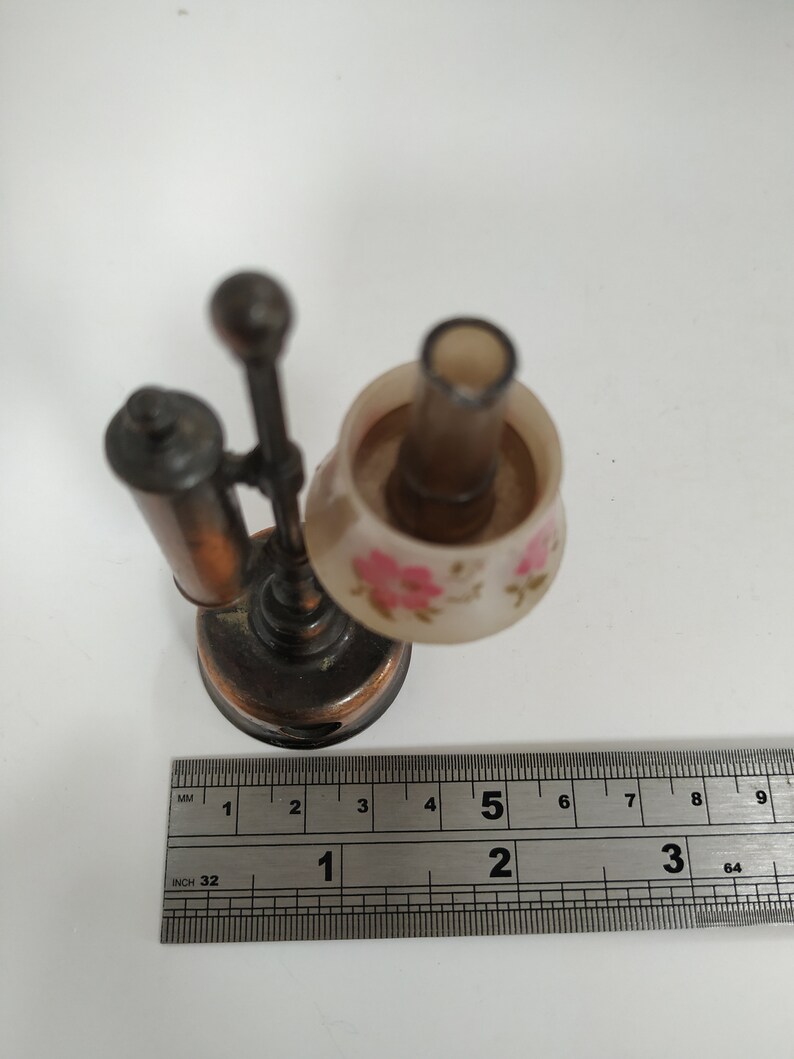 May include: Miniature bronze-colored desk lamp with a floral patterned lampshade. The lamp has a small base and a slender stem. The lampshade is decorated with pink flowers. A ruler is placed next to the lamp.