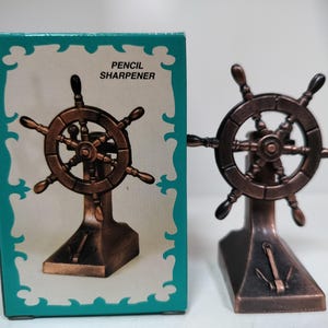 May include: A bronze-coloured pencil sharpener in the shape of a ship's wheel, with a matching box. The box has a teal border and the words "PENCIL SHARPENER". The sharpener has an anchor design on the base.