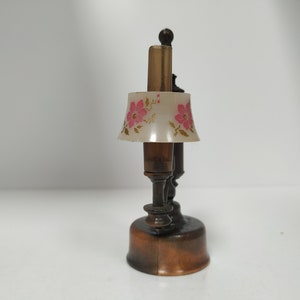 May include: A miniature lamp with a brown base and stem, topped with a lampshade featuring pink floral designs. The lamp has a vintage aesthetic and is likely a decorative item.