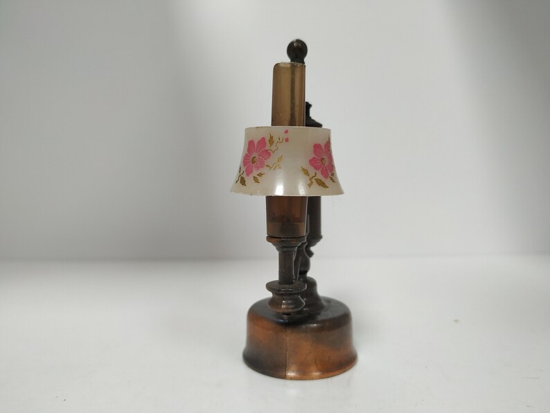 May include: A miniature lamp with a brown base and stem, topped with a lampshade featuring pink floral designs. The lamp has a vintage aesthetic and is likely a decorative item.