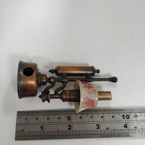 May include: An antique brass and copper-toned lamp with a floral patterned lampshade. The lamp is approximately 4 inches long and features a detailed design. A ruler is placed next to the lamp for scale.