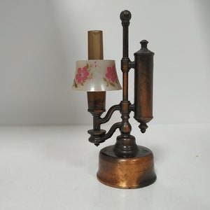 May include: A vintage copper-toned miniature lamp with a small lampshade. The lampshade is white with a pink floral pattern. The lamp has a cylindrical body and a tall, slender design, suggesting a decorative or collectible item.