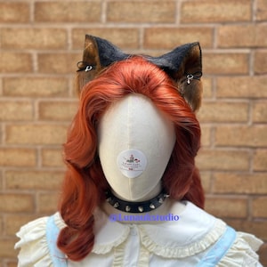 May include: A mannequin head with wavy ginger hair wearing black and brown faux fur animal ears with small black bows and silver charms. A black studded choker is around the neck, and a white collared shirt with light blue trim is visible.