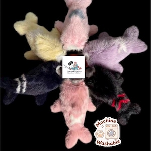 May include: Six plush fish-shaped toys arranged in a circle, featuring soft faux fur in yellow, pink, purple, black with white stripes, solid black, and black with red markings. A central logo reads LunaUKStudio and a sticker says Machine Washable.