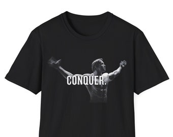 May include: Black t-shirt featuring a black and white image of a bodybuilder with arms raised, and the word "CONQUER" in white block letters. The shirt is made of a soft material and has short sleeves.