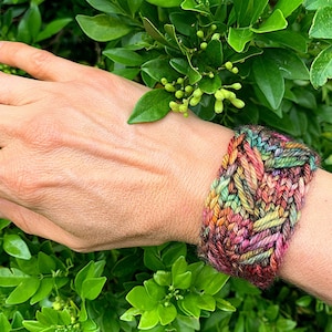 Könnte beinhalten: Ein breites, handgestricktes Armband in verschiedenen Farben, darunter Rot, Orange, Grün und Gelb. Das Armband hat ein strukturiertes, gewebtes Design und wird am Handgelenk getragen. Der Hintergrund ist ein üppiger grüner Busch.