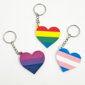 May include: Three heart-shaped keychains with different pride flag designs. The first keychain is rainbow, the second is bisexual, and the third is transgender.