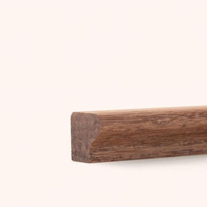 May include: A straight length of medium-brown wood trim featuring a distinct profile with a rounded top edge and a flat base. The wood grain is visible throughout the surface, showing natural variations in color and texture along the length.