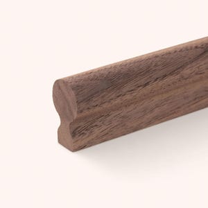 Puede incluir: Una pieza de moldura de madera de nogal marrón oscuro con un perfil superior redondeado y una sección de cuello rebajada. La veta de la madera es visible en toda la superficie, mostrando patrones y texturas naturales a lo largo de la pieza.