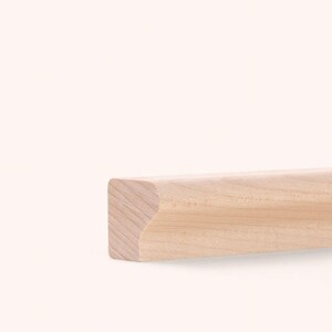 Könnte beinhalten: Eine helle Holzleiste mit einer glatten, natürlich gemaserten Oberfläche. Das Stück hat ein rechteckiges Profil mit einer abgerundeten, geschwungenen Kante an der Oberseite, die die rohe Textur und den hellbraunen Farbton des Holzes zeigt.