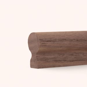Könnte beinhalten: Ein Stück brauner Holzhandlauf mit einem glatten, abgerundeten oberen Profil und einer konturierten, eingekerbten Basis. Das Holz weist eine natürliche Maserung mit sichtbaren dunkelbraunen Streifen auf, die horizontal entlang des Stücks verlaufen.