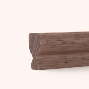 May include: A length of brown wooden molding with a visible grain pattern. The profile features a smooth, curved indentation along the side, creating a distinct shape that runs the entire length of the piece, showcasing the natural wood texture.