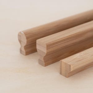 May include: Three wooden picture frame hangers in varying sizes. The hangers are made of light brown bamboo and have a rounded or rectangular profile with a decorative notch. The hangers are arranged diagonally on a light wood surface.