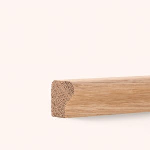 Peut inclure: Une baguette en bois brun clair avec un motif de grain naturel. La pièce présente une section transversale rectangulaire avec un bord arrondi, mettant en valeur le grain d'extrémité texturé sur la gauche et une surface lisse et allongée.