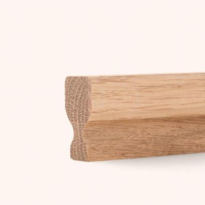 May include: A length of unfinished light brown oak wood trim featuring a distinct profile with a rounded top edge, a concave middle section, and a flat bottom edge, showcasing the natural grain patterns and texture of the solid wood material.
