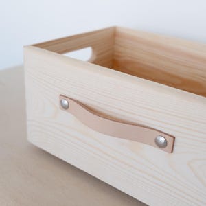 Könnte beinhalten: Eine Aufbewahrungsbox aus hellem Holz mit einem Ledergriff und Metallnieten. Die Box ist leer und bereit, mit Gegenständen gefüllt zu werden.