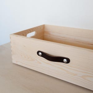 Puede incluir: Una caja de almacenamiento de madera con una manija de cuero marrón y remaches de metal plateado. La caja está hecha de madera clara y tiene un acabado natural.
