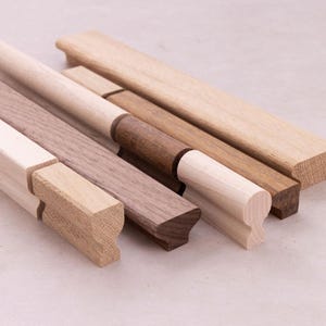 May include: A collection of linear wooden cabinet pulls featuring varied wood grains and tones, including light maple and dark walnut. Each handle has a distinct profile with a rounded top edge and a recessed grip, showcasing natural wood textures.