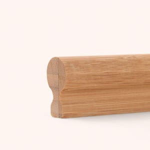 Può includere: Un corrimano in legno marrone chiaro con un profilo superiore liscio e arrotondato e una sezione laterale sagomata e rientrante. La venatura del legno è visibile lungo tutta la lunghezza del pezzo, mostrando una finitura naturale calda.