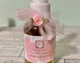 May include: A clear glass spray bottle filled with a brown liquid, labeled "Arts in Suds Cocoa Butter Cashmere Moisturizing Body Spray." The bottle is adorned with a pink bow and a small pink rose. The bottle contains 2 fl oz.