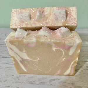 May include: Two bars of handmade soap. The soap has a light beige base with white and pink swirls. The top of each bar is decorated with white, geometric shapes and pink sprinkles. The soap is a rectangular shape.