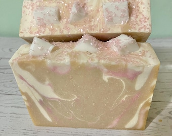 May include: Two bars of handmade soap. The soap has a light beige base with white and pink swirls. The top of each bar is decorated with white, geometric shapes and pink sprinkles. The soap is a rectangular shape.