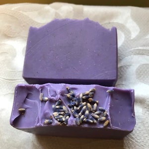 May include: Two bars of lavender soap, one with dried lavender buds on top. The soap is a light purple color.