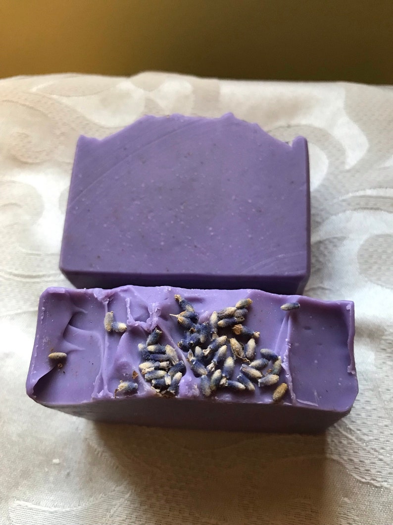 May include: Two bars of lavender soap, one with dried lavender buds on top. The soap is a light purple color.