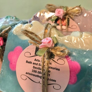 May include: A blue and white soap bar with pink flower embellishments. The soap is wrapped in clear plastic and tied with twine. A pink and white label with the text "Arts in Suds, Bath and Body Luxuries, Seville, Ohio, 330-388-8111, artsinsuds@y.com" is attached to the soap.