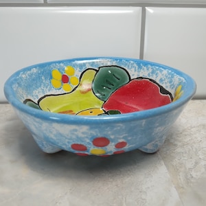 May include: A small, blue ceramic bowl with three feet. The bowl is decorated with hand-painted yellow, green, and red fruit and flowers.
