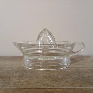May include: A clear glass citrus juicer with a reamer and a bowl with a handle. The bowl has horizontal ridges and measurement markings. The reamer is cone-shaped with vertical ridges. The juicer sits on a wooden surface.