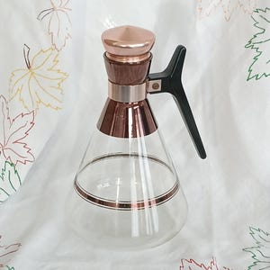 May include: A clear glass carafe with a copper-colored lid, trim, and a handle. The carafe has a conical shape with a copper-colored band around the middle. The handle is black. The carafe is likely for serving beverages.