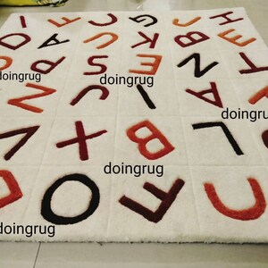 K&ouml;nnte beinhalten: Wei&szlig;er Teppich mit braunen, roten und schwarzen Buchstaben, die darauf gestickt sind. Die Buchstaben sind in zuf&auml;lliger Reihenfolge angeordnet und buchstabieren mehrmals das Wort "doingrug".