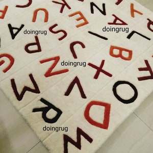 K&ouml;nnte beinhalten: Ein wei&szlig;er Teppich mit den Buchstaben des Alphabets, die in verschiedenen Farben, darunter Rot, Braun und Schwarz, gestickt sind. Die Buchstaben sind in einem zuf&auml;lligen Muster angeordnet. Der Teppich besteht aus einem weichen, pl&uuml;schigen Material.