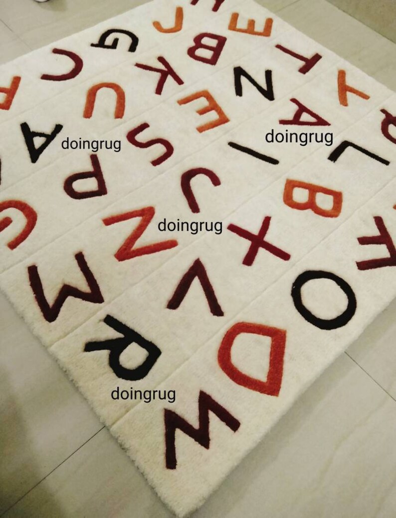 K&ouml;nnte beinhalten: Ein wei&szlig;er Teppich mit den Buchstaben des Alphabets, die in verschiedenen Farben, darunter Rot, Braun und Schwarz, gestickt sind. Die Buchstaben sind in einem zuf&auml;lligen Muster angeordnet. Der Teppich besteht aus einem weichen, pl&uuml;schigen Material.