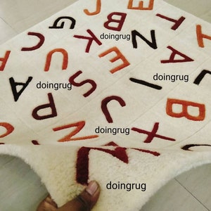 K&ouml;nnte beinhalten: Wei&szlig;er Alphabet-Teppich mit in Orange, Rot und Braun gestickten Buchstaben. Der Teppich ist in der Mitte gefaltet und zeigt die wei&szlig;e R&uuml;ckseite.