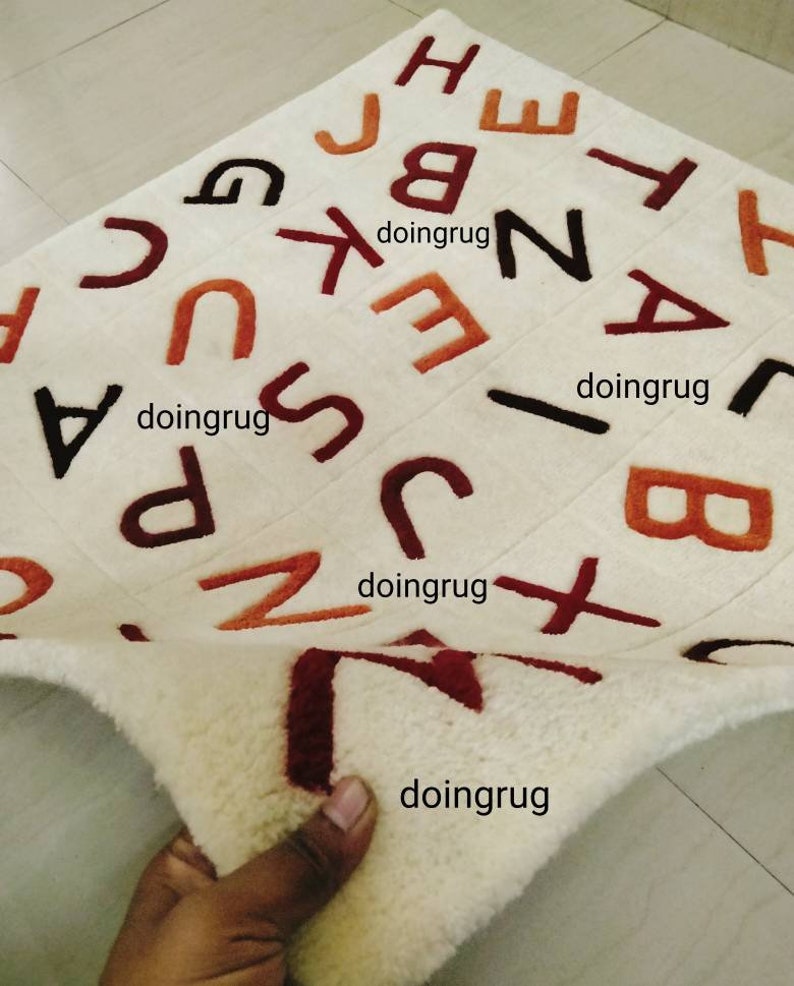 K&ouml;nnte beinhalten: Wei&szlig;er Alphabet-Teppich mit in Orange, Rot und Braun gestickten Buchstaben. Der Teppich ist in der Mitte gefaltet und zeigt die wei&szlig;e R&uuml;ckseite.