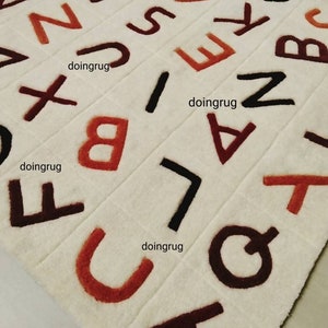 K&ouml;nnte beinhalten: Ein wei&szlig;er Teppich mit braunen, schwarzen und orangen Buchstaben des Alphabets. Die Buchstaben sind in verschiedenen Gr&ouml;&szlig;en und in einem zuf&auml;lligen Muster angeordnet. Der Teppich besteht aus einem weichen, pl&uuml;schigen Material.