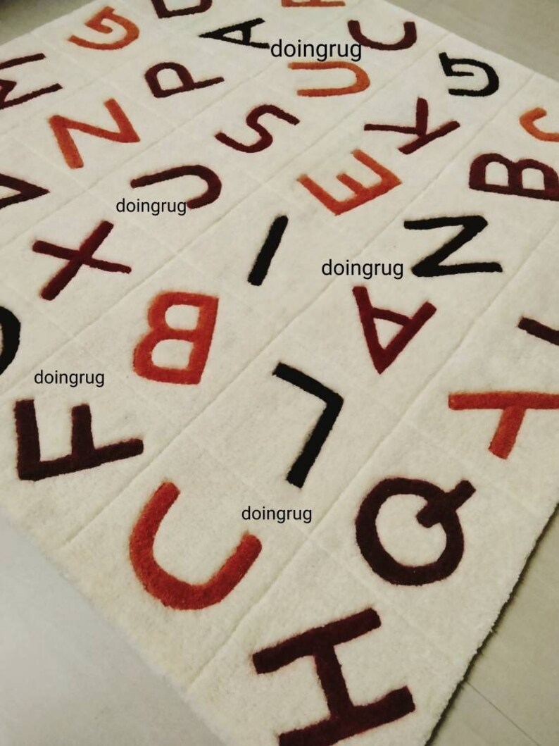 K&ouml;nnte beinhalten: Ein wei&szlig;er Teppich mit braunen, schwarzen und orangen Buchstaben des Alphabets. Die Buchstaben sind in verschiedenen Gr&ouml;&szlig;en und in einem zuf&auml;lligen Muster angeordnet. Der Teppich besteht aus einem weichen, pl&uuml;schigen Material.