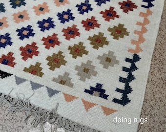 May include: A white rug with a geometric pattern in shades of blue, red, green, and brown. The rug has a fringe border and the text "doing rugs" is visible in the bottom right corner.