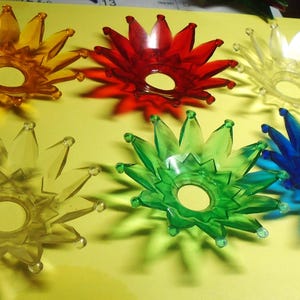 May include: A collection of six translucent, star-shaped glass or plastic bobeches arranged in two rows. The colors include amber, red, clear, light green, and blue. Each piece features a central circular opening and pointed, petal-like edges.
