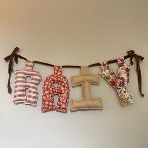 May include: A fabric banner spelling out the word "BABY" in individual letter shapes. The letters are made of various patterned fabrics, including red and white stripes, floral prints, and solid beige. The banner is strung on a brown cord with brown ribbon ties.