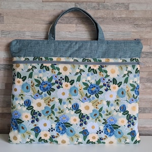 Può includere: Una borsa tote floreale con parte superiore e manici in denim blu. La borsa presenta un motivo di fiori blu, bianchi e gialli su sfondo azzurro. Ha una chiusura a cerniera ed è progettata per trasportare oggetti.