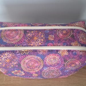 Può includere: Un beauty case rettangolare viola con un motivo mandala ripetuto in tonalità rosa, oro e blu. La borsa ha due cerniere con tiretti color crema. La borsa è appoggiata su una superficie di legno.